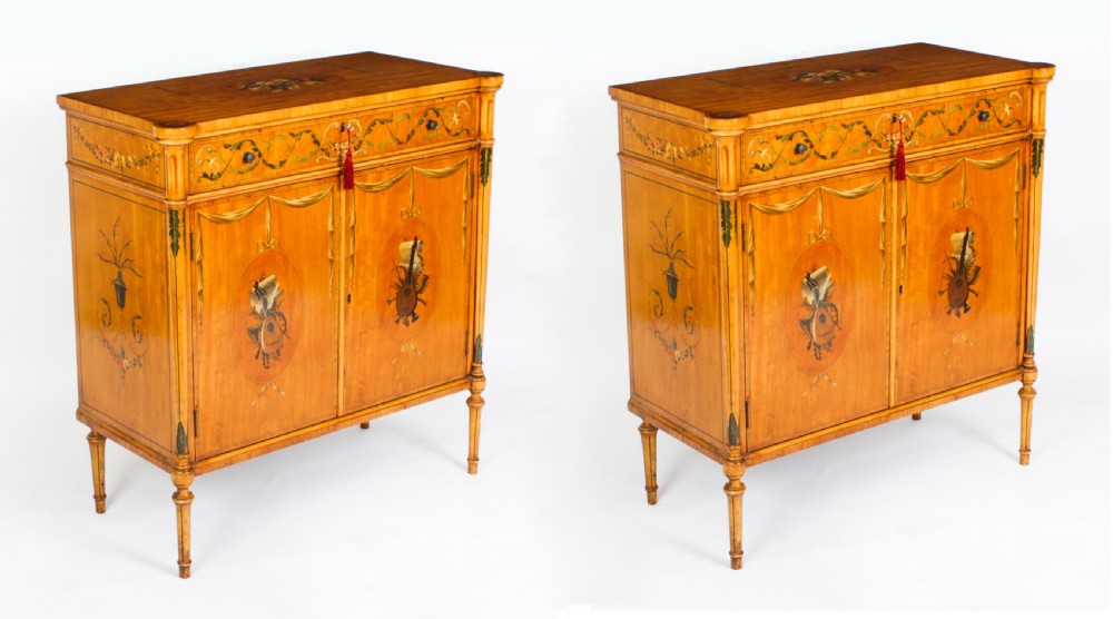 A pair of 19th-century Adam Revival painted satinwood side cabinets featuring painted oval musical medallions, draped garlands, single top drawers, double cupboards, and turned tapered legs.