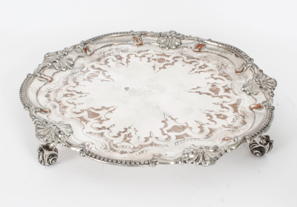 A Georgian Old Sheffield plate silver-plated salver featuring chased scroll engravings, shell motifs on a beaded rim, and resting on short feet.