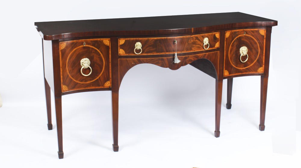 An 18th-century George III flame mahogany serpentine sideboard featuring inlaid circular panel doors, brass lion-mask ring handles, and tapered square legs terminating in spade feet.