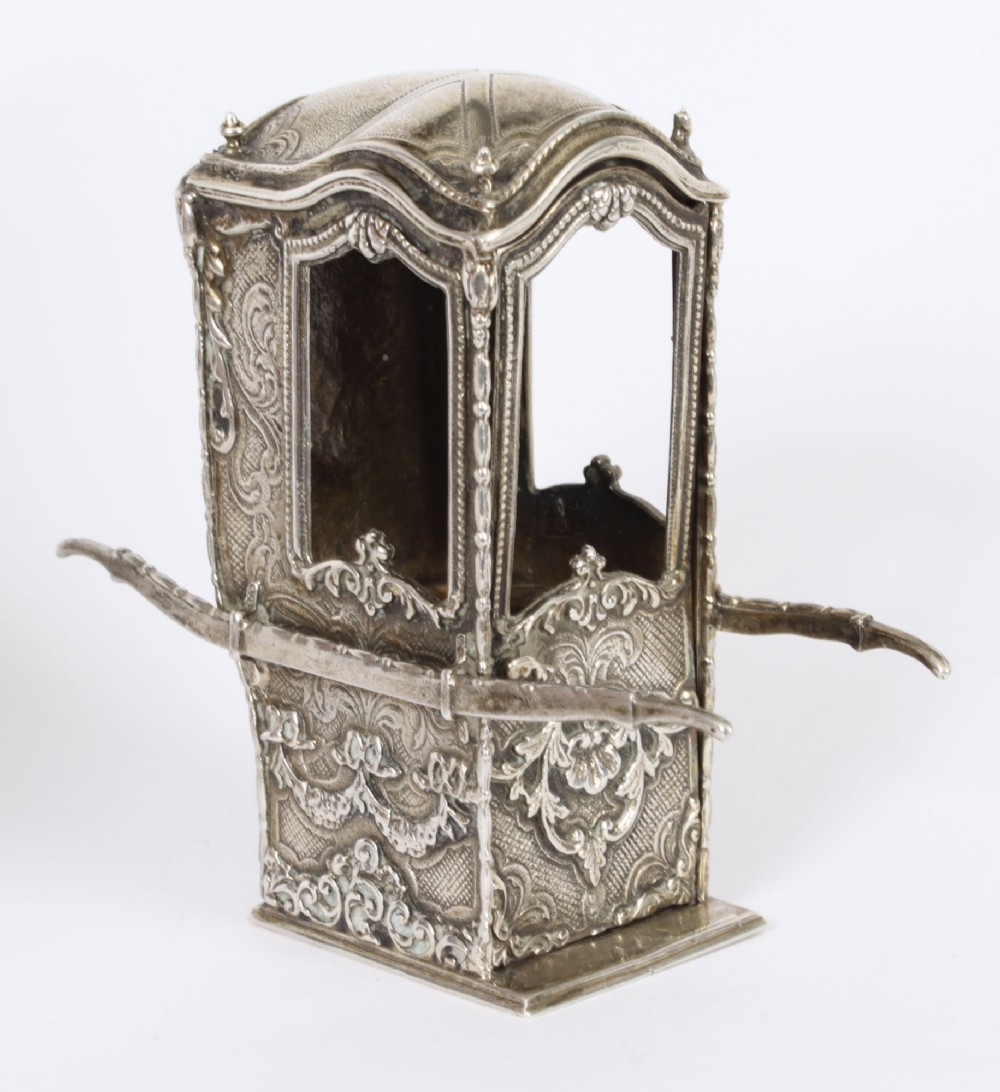 A detailed 19th-century silver miniature sedan chair featuring ornate Rococo floral chasing, side carrying poles, a shaped roof, and window openings.