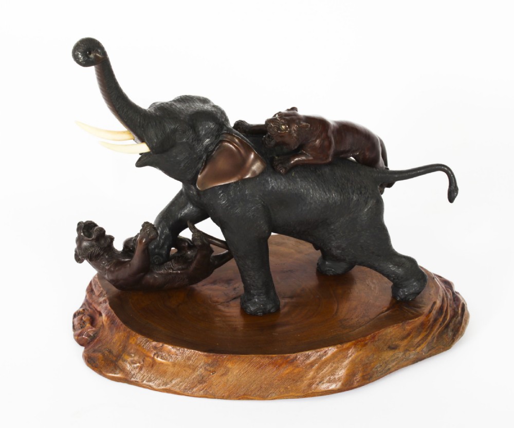 A dark patinated bronze sculpture of an elephant defending against two tigers, mounted on a polished natural wood base.