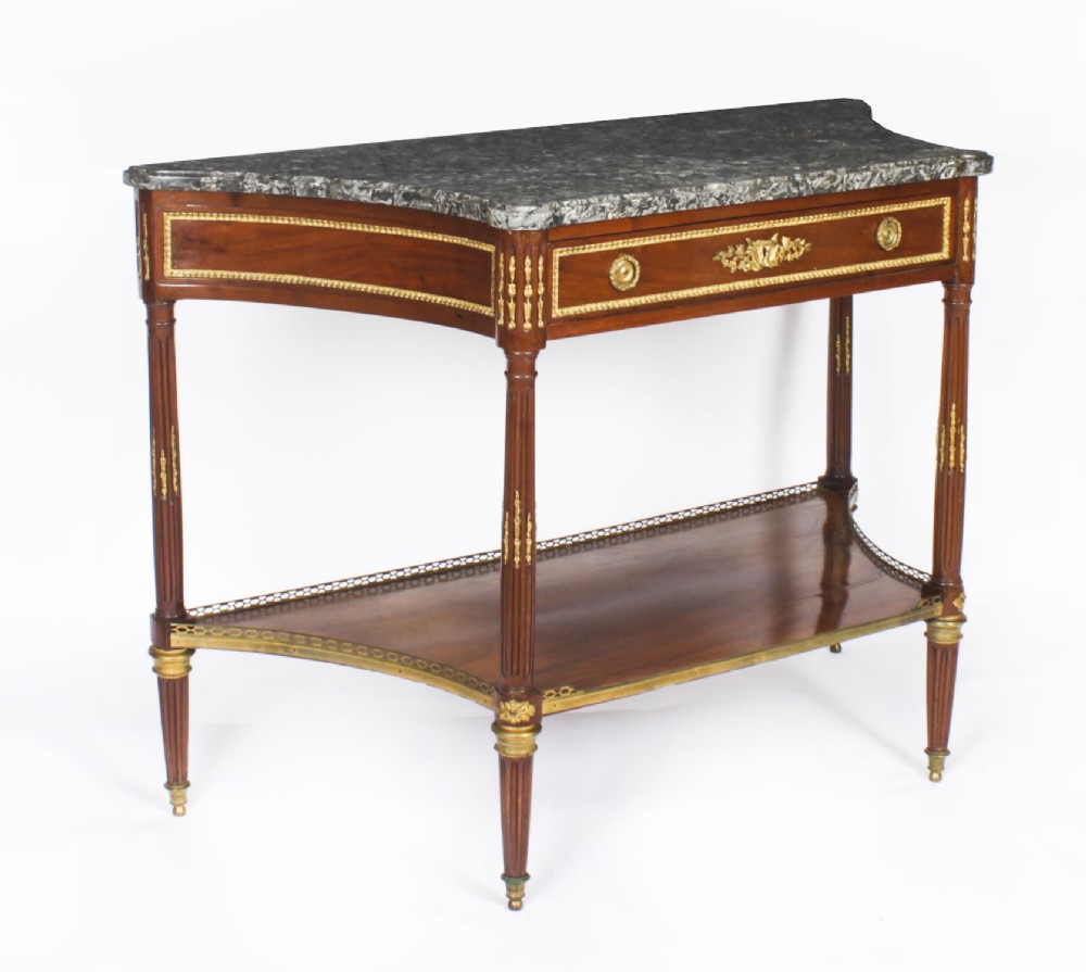 An antique French Louis XVI Revival mahogany console table featuring a grey marble top, gilt-bronze mounts, fluted tapered legs, and a lower tier shelf.