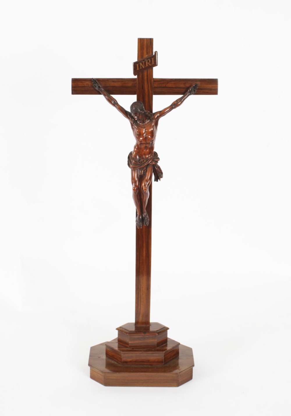 A carved wooden Corpus Christi mounted on a dark olive wood cross with an INRI scroll, supported by a tiered hexagonal wooden base.
