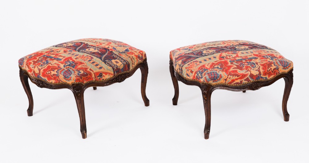 A pair of 19th-century French walnut footstools featuring carved cabriole legs and patterned red floral tapestry upholstery secured with brass studs.