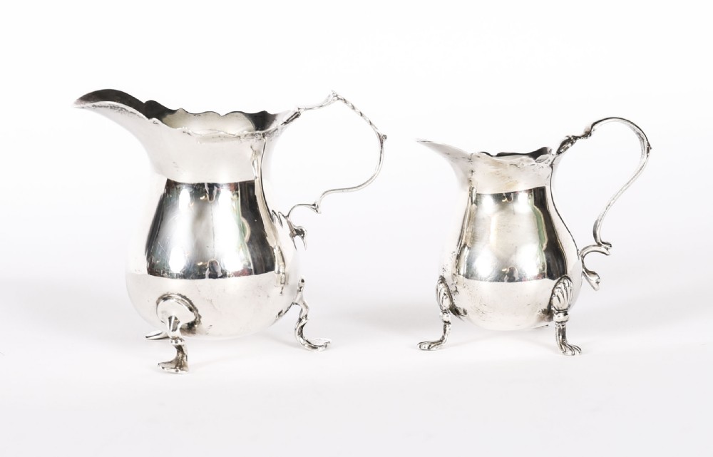 A graduating pair of Victorian sterling silver cream jugs featuring scalloped rims, scroll handles, and tripod legs.