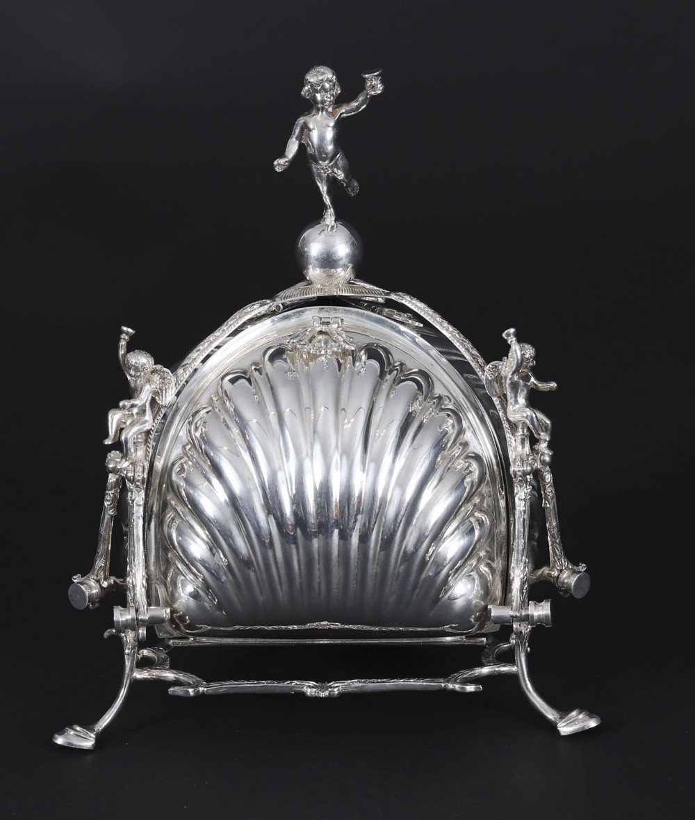 An antique silver-plated folding biscuit box featuring scalloped shell compartments, a top cherub finial, and decorative figurative side supports.