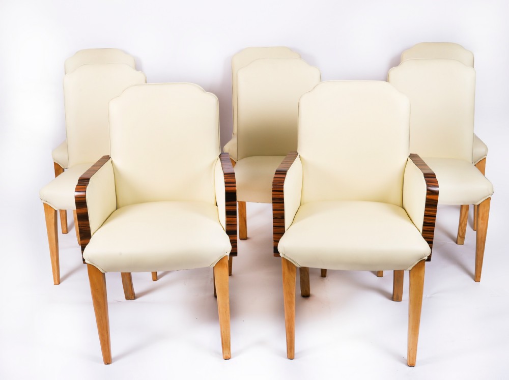 A set of eight Art Deco dining chairs featuring cream leather upholstery, curved cloud-shaped backrests, and tapered timber legs, including two carvers with dark veneered armrests.