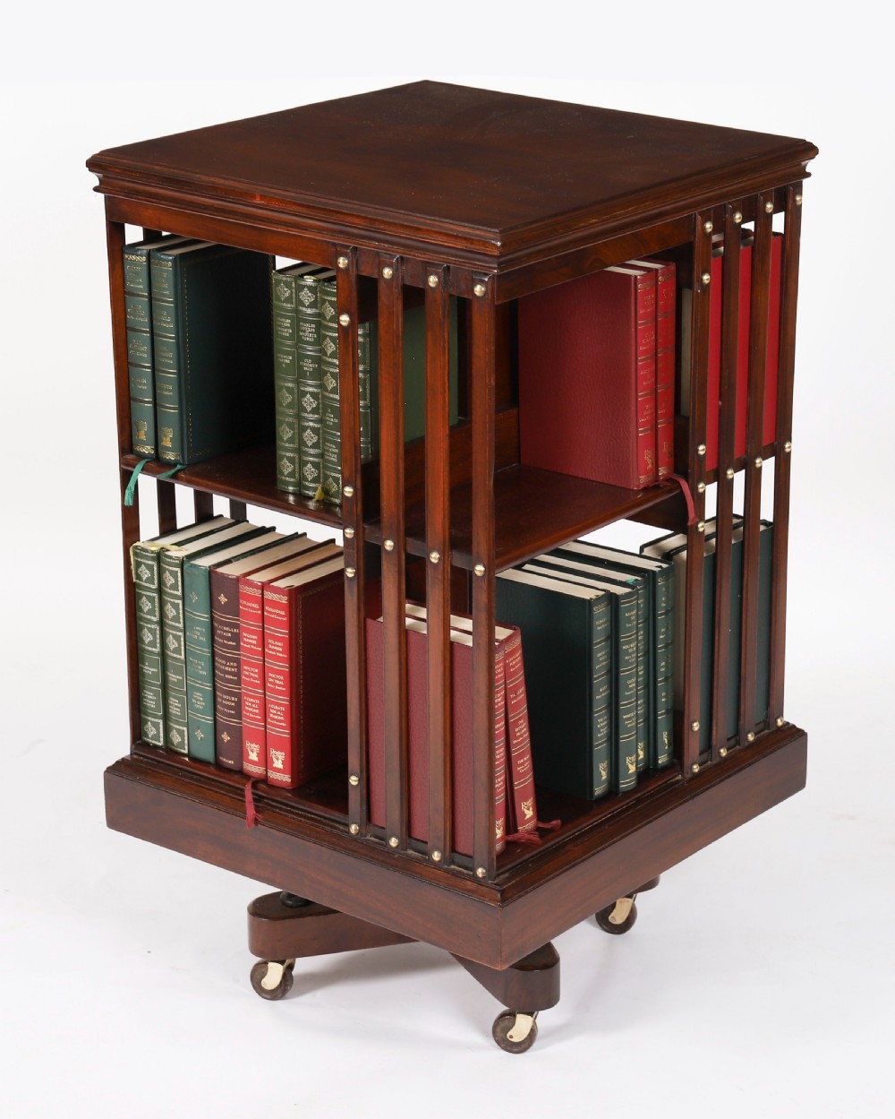 A two-tier dark mahogany revolving bookcase featuring vertical slatted sides with brass stud detailing, resting on a quadform base with castors.