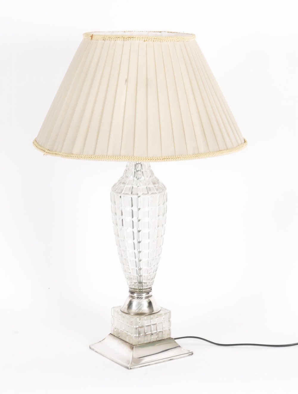 An Art Deco table lamp featuring a faceted clear cut crystal stem, a square metal pedestal base, and a cream pleated fabric lampshade.