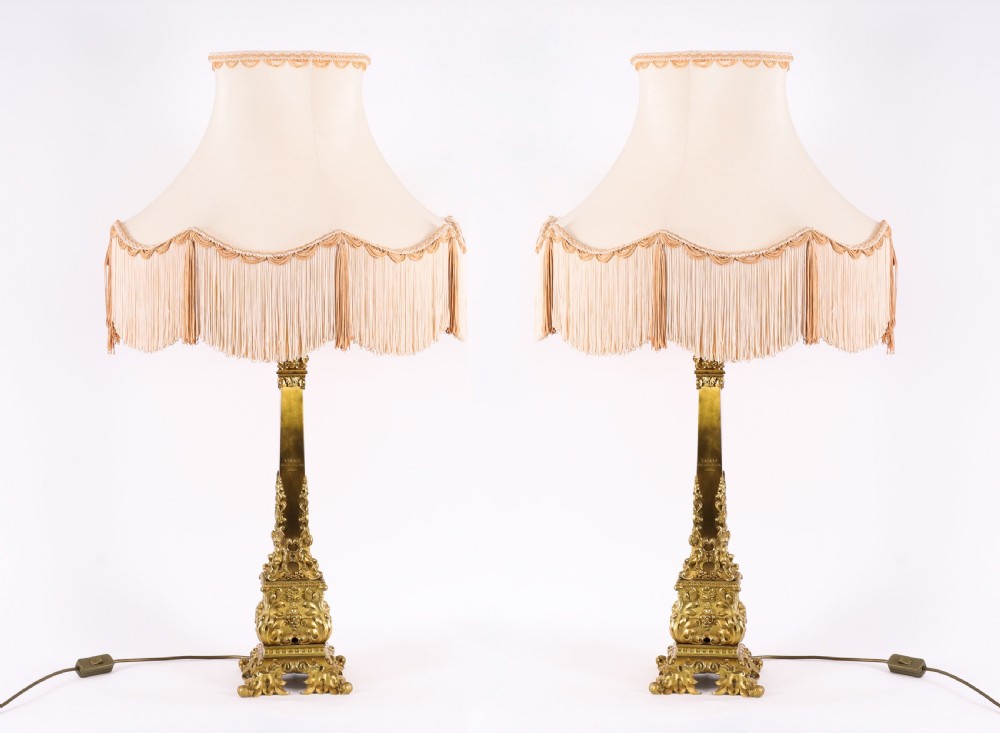 A pair of 19th-century French ormolu table lamps featuring cast gilt bronze bases and cream flared fabric shades with tassel fringe trim.