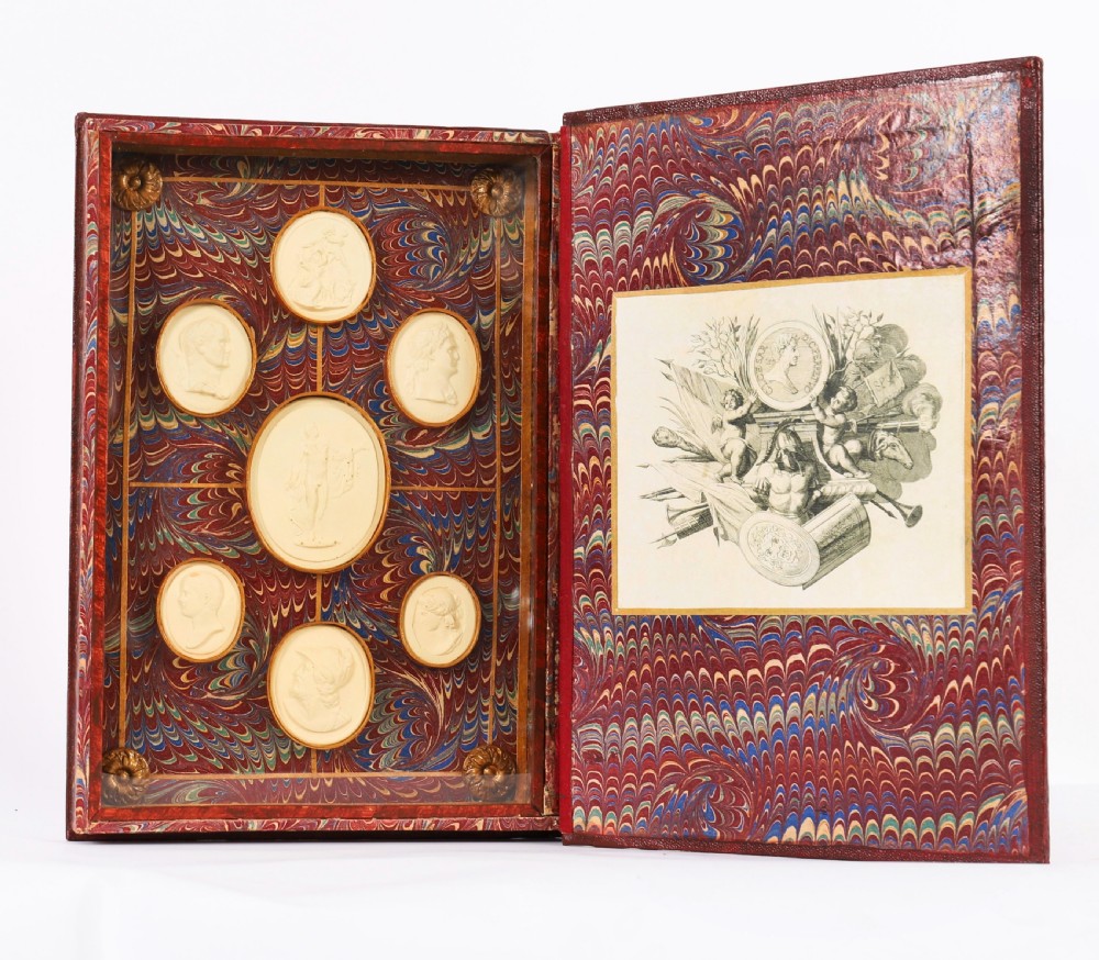 An open red marbled book-form box displaying seven oval cream plaster intaglios behind glass alongside an engraved frontispiece. View II