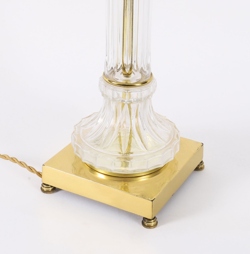 A pair of Art Deco table lamps featuring fluted clear glass column stems on square gilded metal bases with arched cream lampshades. View X