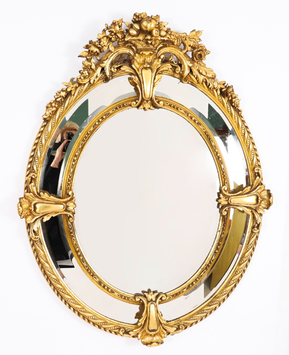 An ornate nineteenth-century oval giltwood cushion mirror featuring carved floral cresting, acanthus leaf accents, and surround mirror paneling.