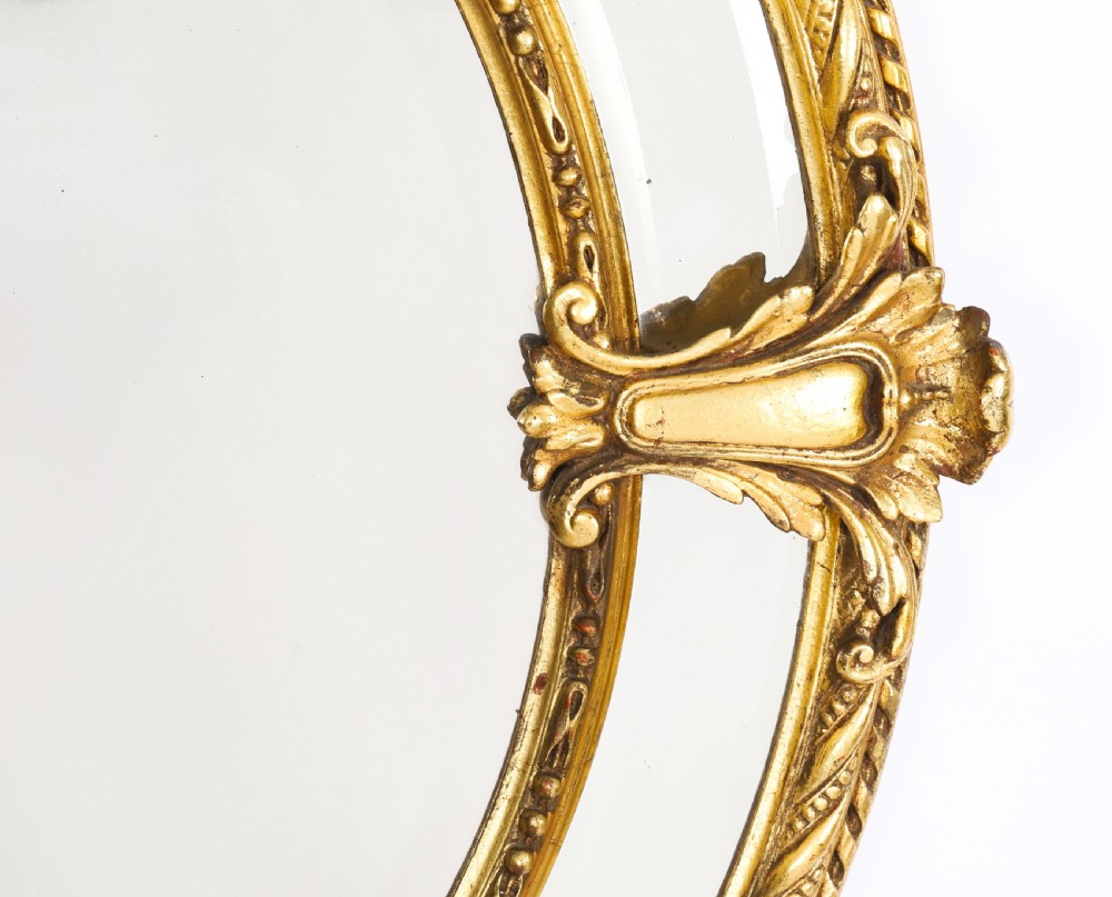 An ornate nineteenth-century oval giltwood cushion mirror featuring carved floral cresting, acanthus leaf accents, and surround mirror paneling. View V