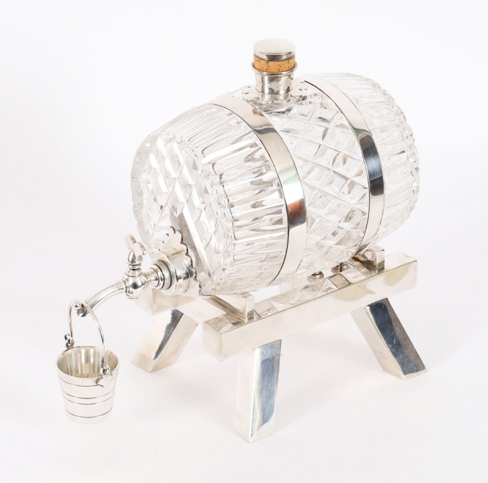 A nineteenth-century cut crystal barrel decanter with silver-plated hoops, tap, and miniature bucket accessory resting on a four-legged silver stand.
