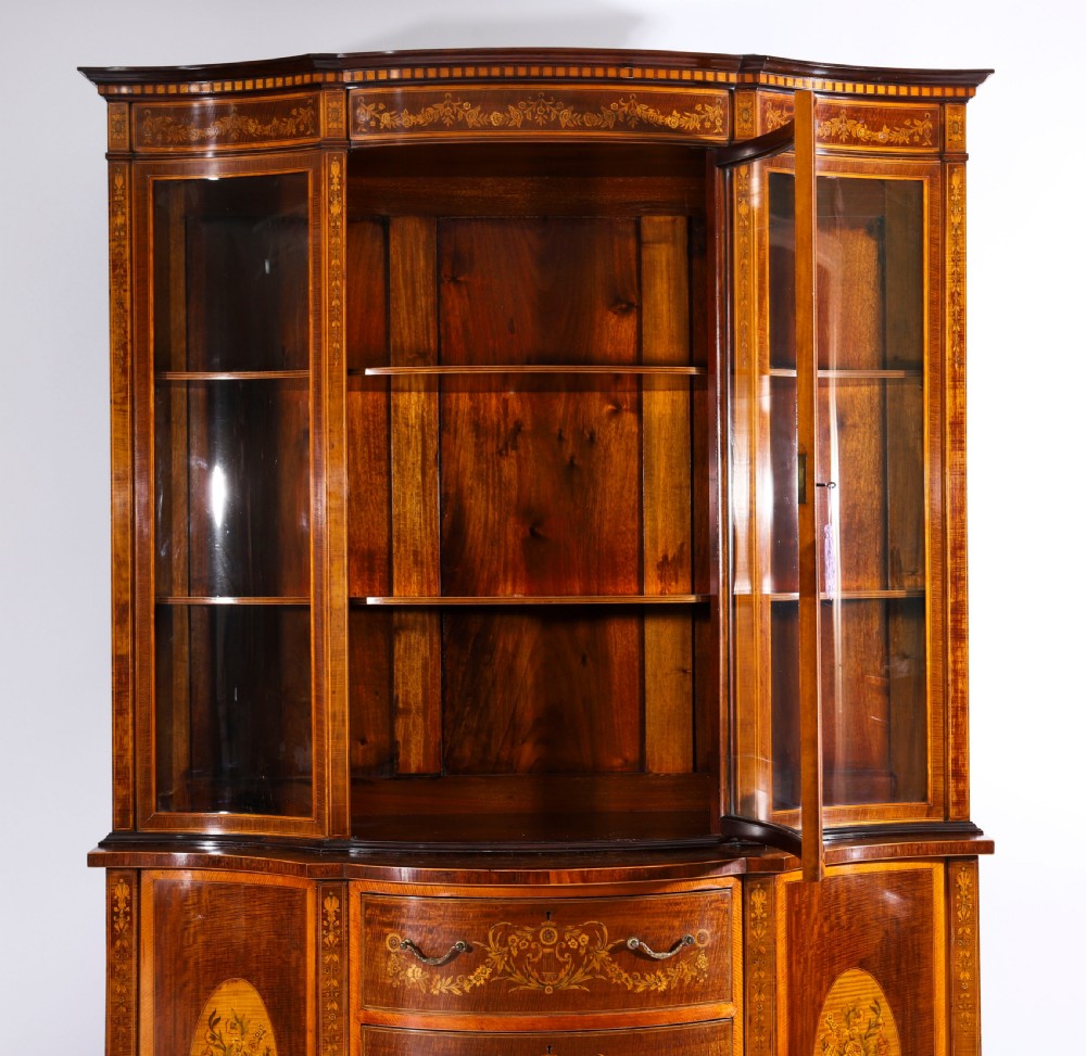 A 19th-century mahogany serpentine display cabinet featuring intricate floral marquetry inlays, three glazed upper doors, three central drawers, and tapered legs. View IV