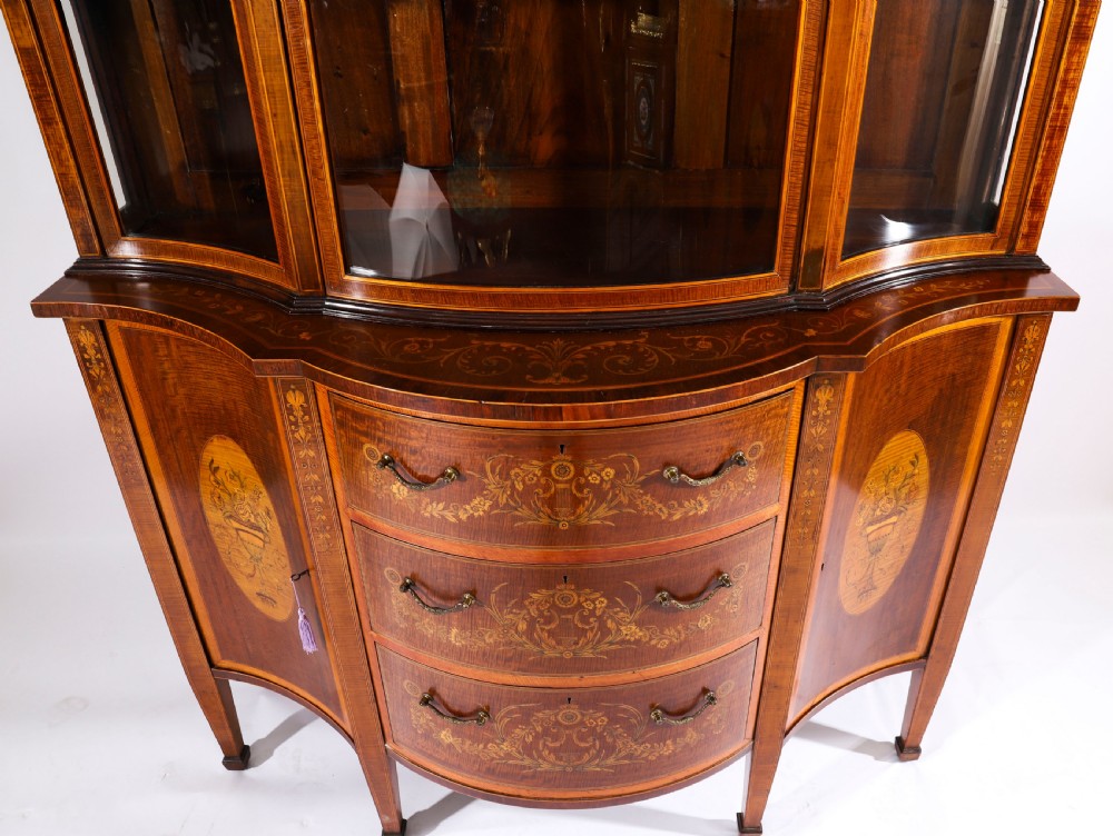 A 19th-century mahogany serpentine display cabinet featuring intricate floral marquetry inlays, three glazed upper doors, three central drawers, and tapered legs. View XV