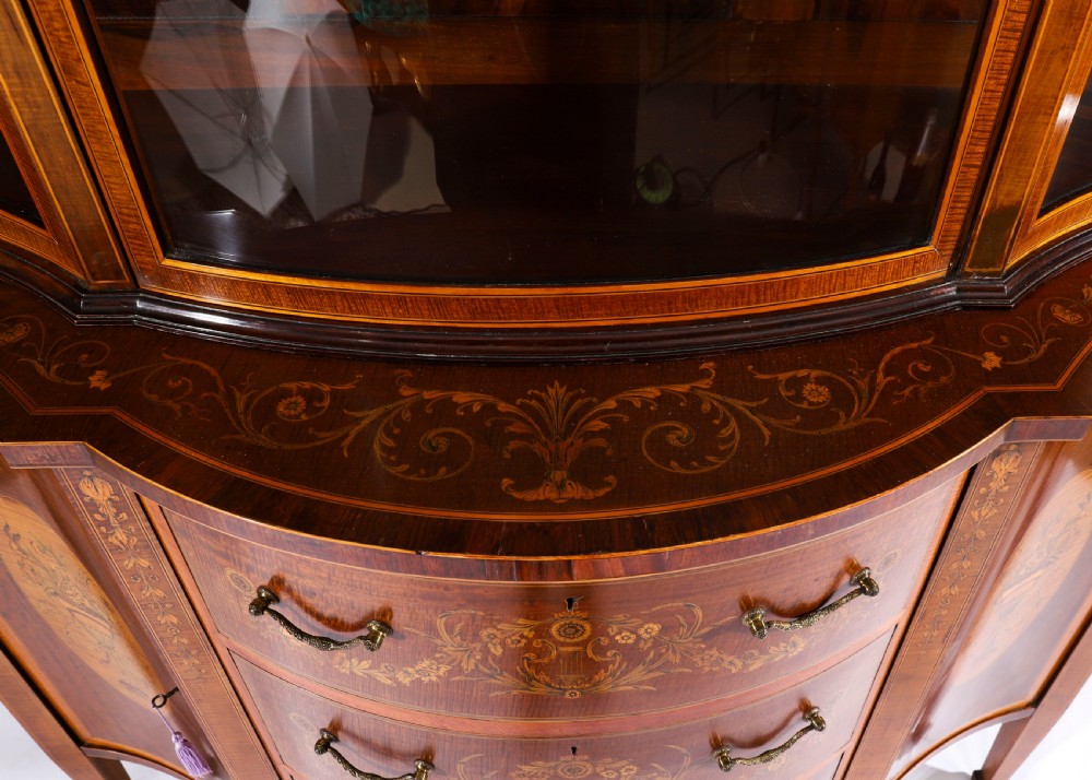 A 19th-century mahogany serpentine display cabinet featuring intricate floral marquetry inlays, three glazed upper doors, three central drawers, and tapered legs. View XVI
