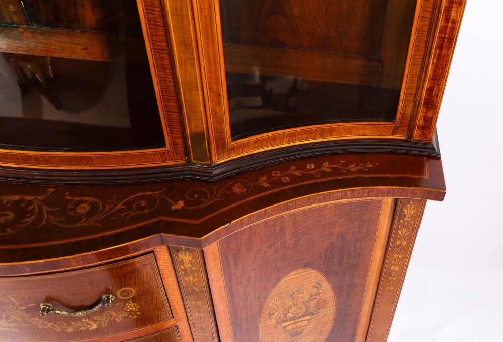 A 19th-century mahogany serpentine display cabinet featuring intricate floral marquetry inlays, three glazed upper doors, three central drawers, and tapered legs. View XVII