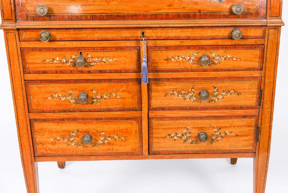 A late Victorian Sheraton Revival satinwood cylinder bureau featuring hand-painted floral swags, a central figural scene, and multiple small drawers above tapered legs. View X
