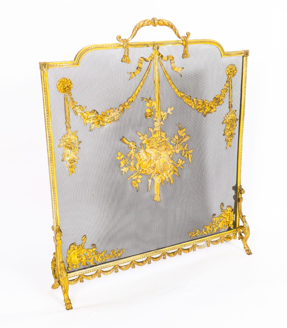 A 19th-century French gilt bronze fire screen featuring a woven mesh panel decorated with central musical trophy motifs, floral garlands, and ribbon swags.
