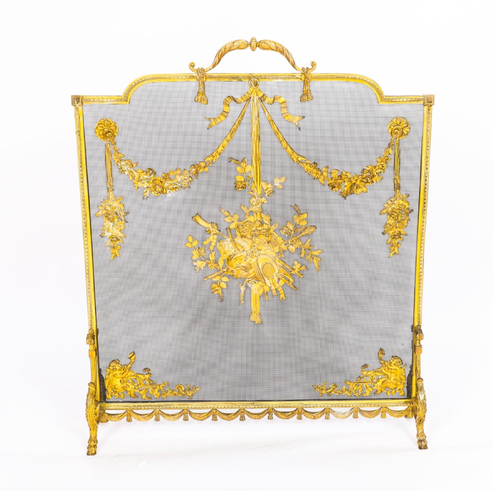 A 19th-century French gilt bronze fire screen featuring a woven mesh panel decorated with central musical trophy motifs, floral garlands, and ribbon swags. View V