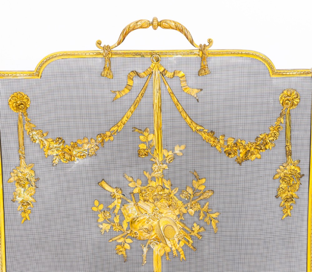 A 19th-century French gilt bronze fire screen featuring a woven mesh panel decorated with central musical trophy motifs, floral garlands, and ribbon swags. View X