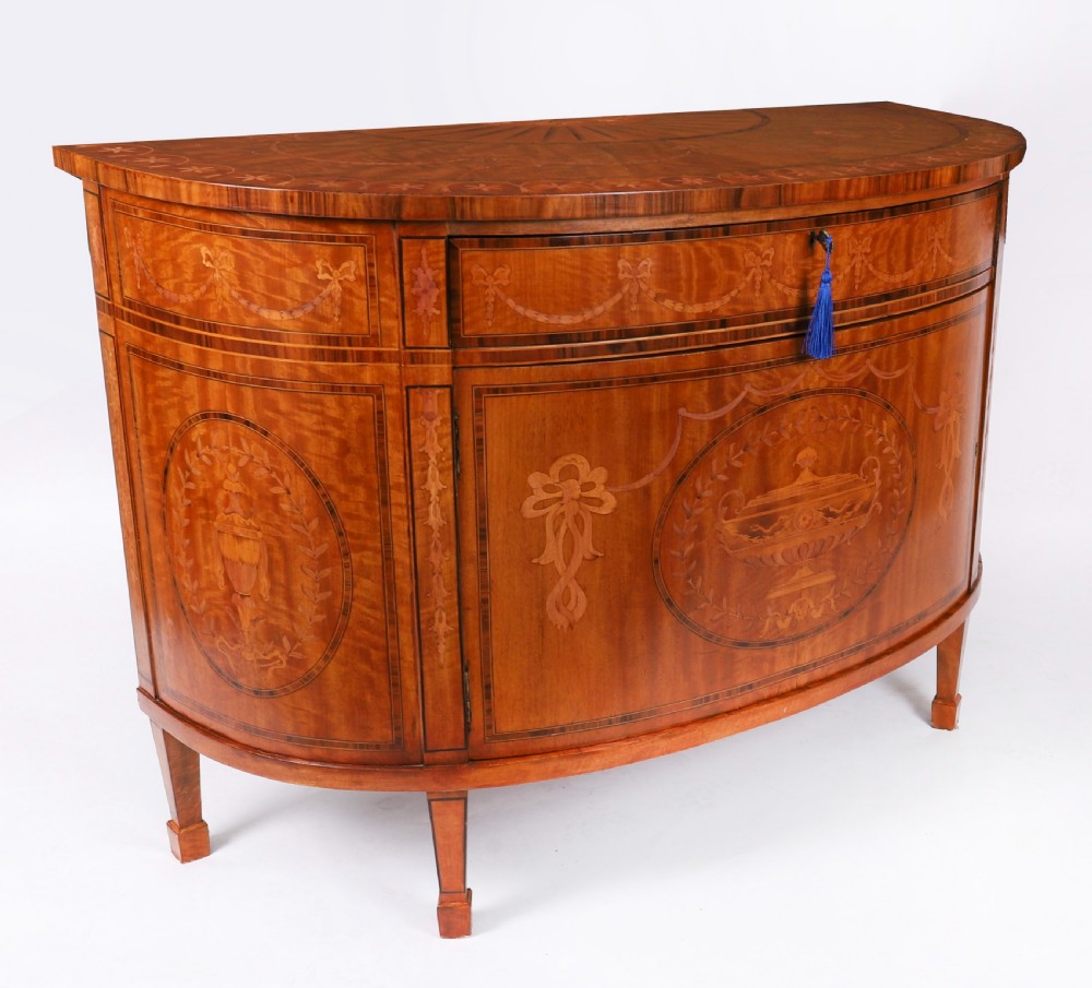 A semicircular antique satinwood demilune cabinet featuring urn and swag marquetry inlays, a top drawer with a blue tassel key, and slender tapered legs.