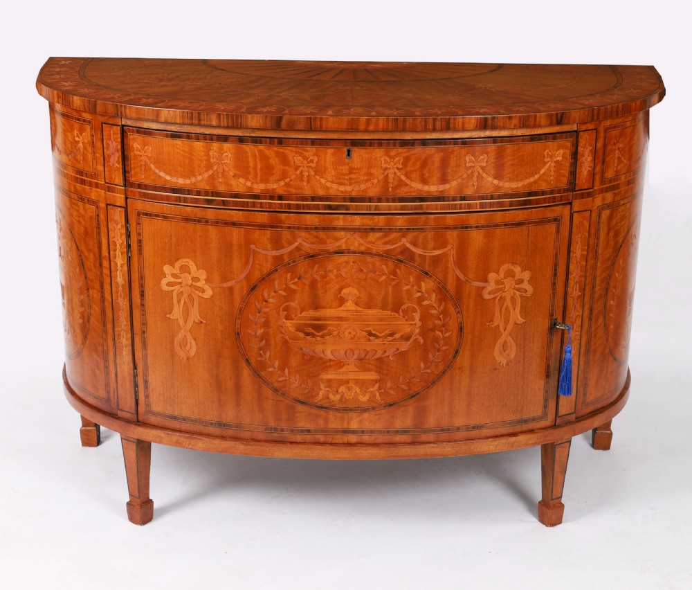 A semicircular antique satinwood demilune cabinet featuring urn and swag marquetry inlays, a top drawer with a blue tassel key, and slender tapered legs. View II