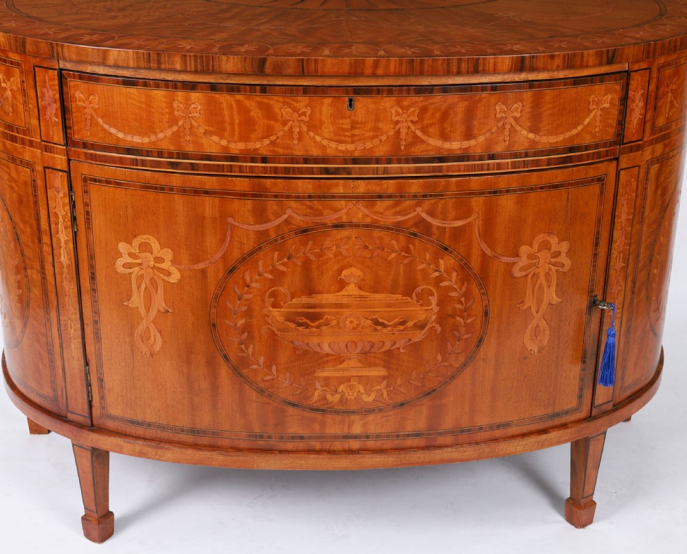 A semicircular antique satinwood demilune cabinet featuring urn and swag marquetry inlays, a top drawer with a blue tassel key, and slender tapered legs. View IV