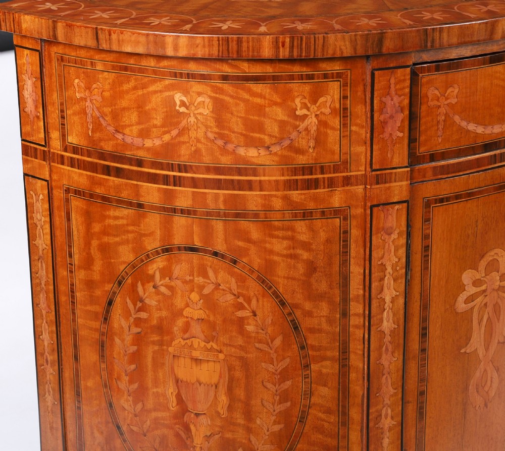 A semicircular antique satinwood demilune cabinet featuring urn and swag marquetry inlays, a top drawer with a blue tassel key, and slender tapered legs. View VII