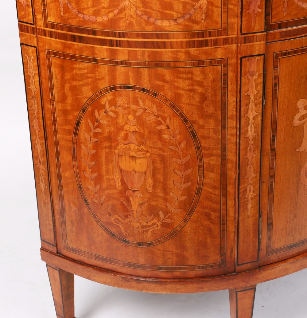 A semicircular antique satinwood demilune cabinet featuring urn and swag marquetry inlays, a top drawer with a blue tassel key, and slender tapered legs. View IX