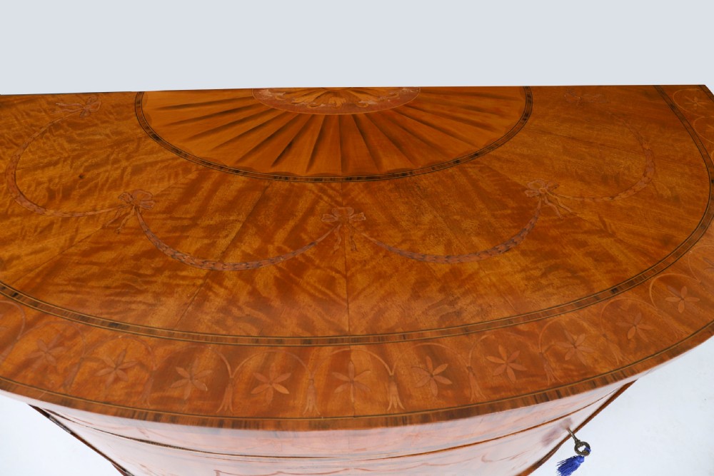 A semicircular antique satinwood demilune cabinet featuring urn and swag marquetry inlays, a top drawer with a blue tassel key, and slender tapered legs. View X
