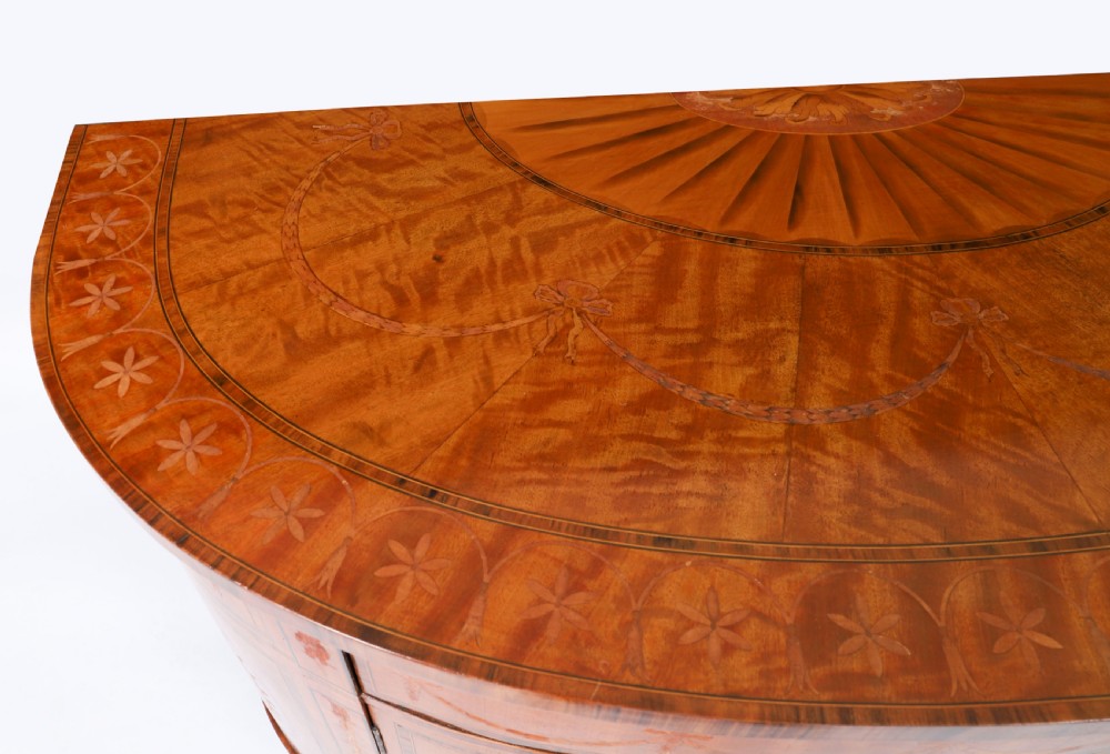 A semicircular antique satinwood demilune cabinet featuring urn and swag marquetry inlays, a top drawer with a blue tassel key, and slender tapered legs. View XI