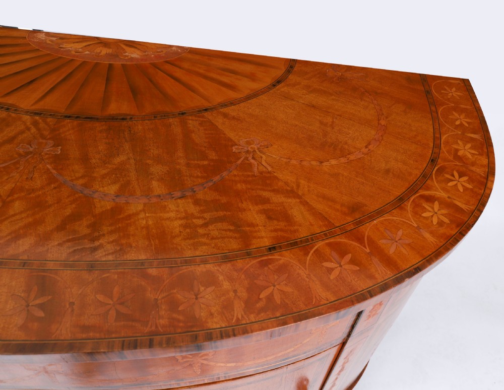 A semicircular antique satinwood demilune cabinet featuring urn and swag marquetry inlays, a top drawer with a blue tassel key, and slender tapered legs. View XIII