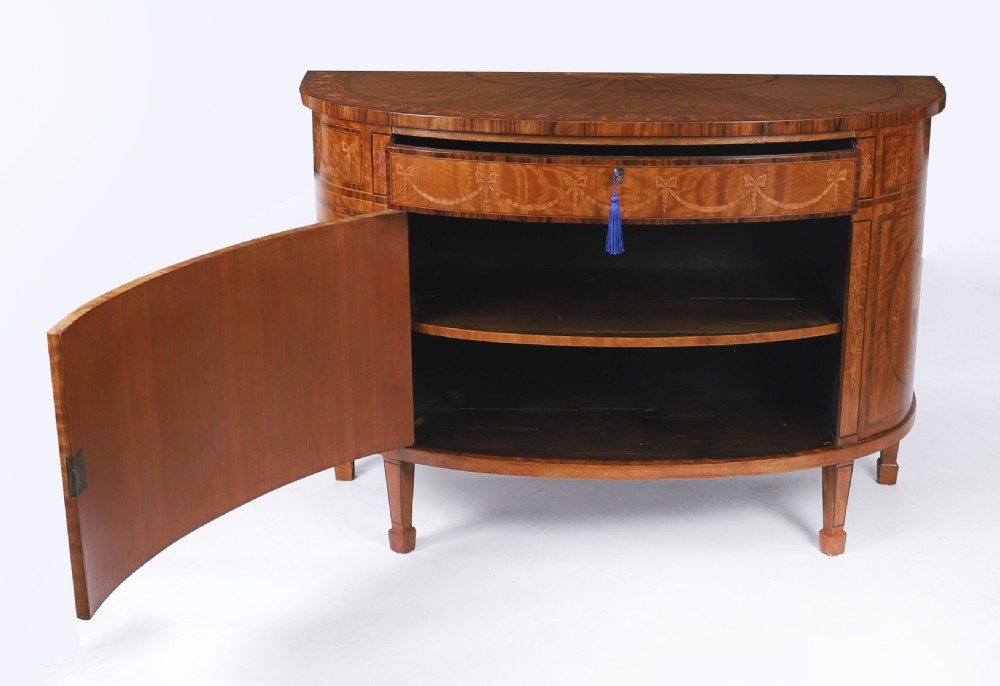 A semicircular antique satinwood demilune cabinet featuring urn and swag marquetry inlays, a top drawer with a blue tassel key, and slender tapered legs. View XIV