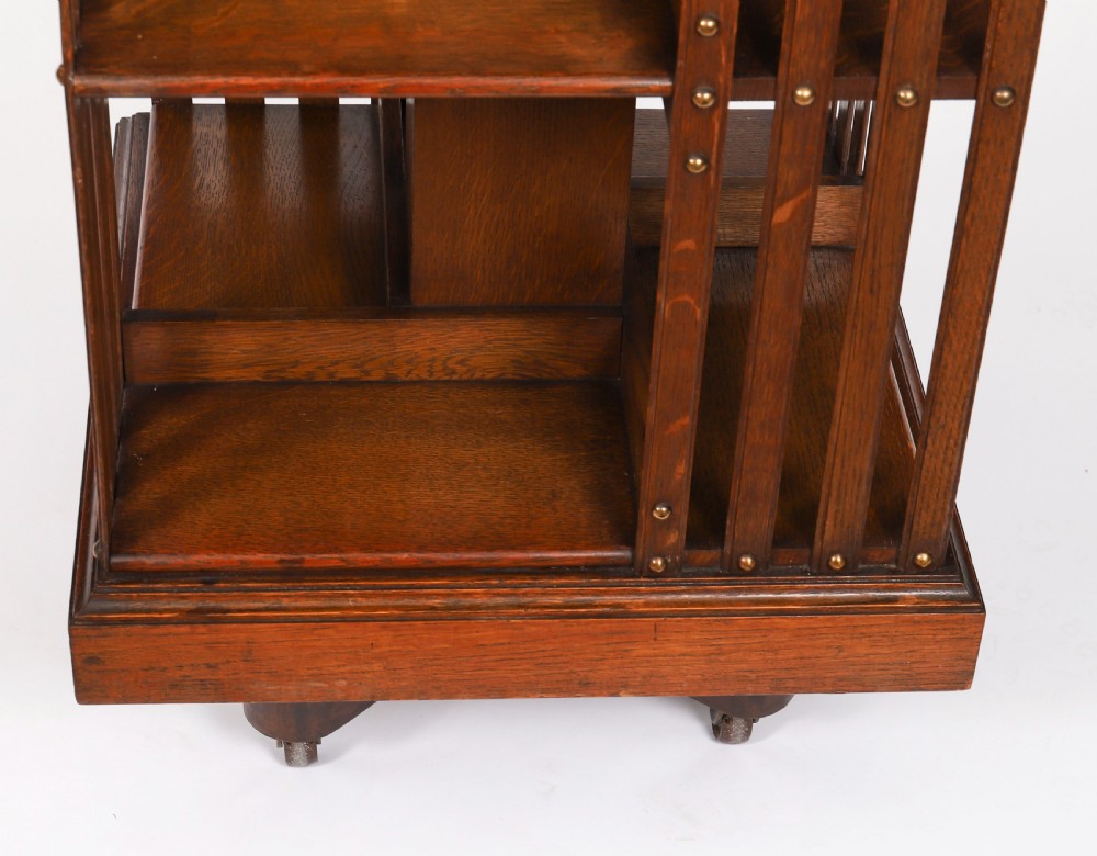 A two-tier Victorian dark oak revolving bookcase with vertical side slats and brass studs, raised on a quadruped base with metal castors. View X