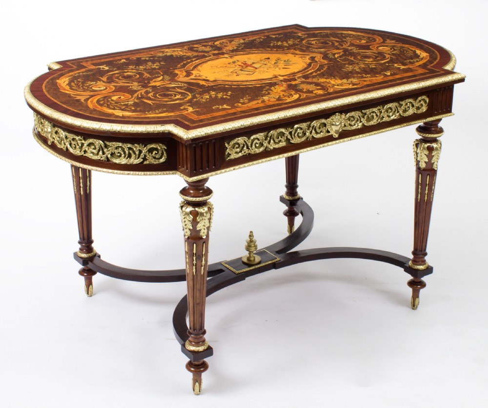 An antique French bureau plat writing table featuring detailed floral marquetry inlays, decorative gilt-bronze ormolu mounts, turned legs, and an X-frame stretcher.