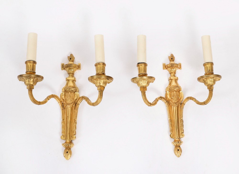 A pair of antique French Louis XVI style two-branch ormolu wall lights with fluted backplates and cream candle sleeves.