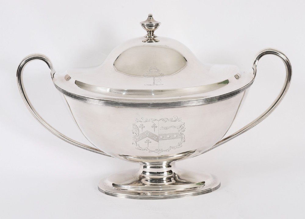 A George III sterling silver tureen with two looped handles, a domed lid, a pedestal base, and an engraved heraldic crest.