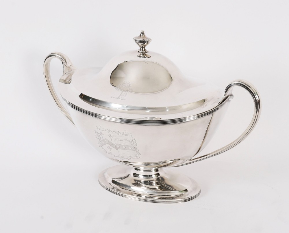 A George III sterling silver tureen with two looped handles, a domed lid, a pedestal base, and an engraved heraldic crest. View XV