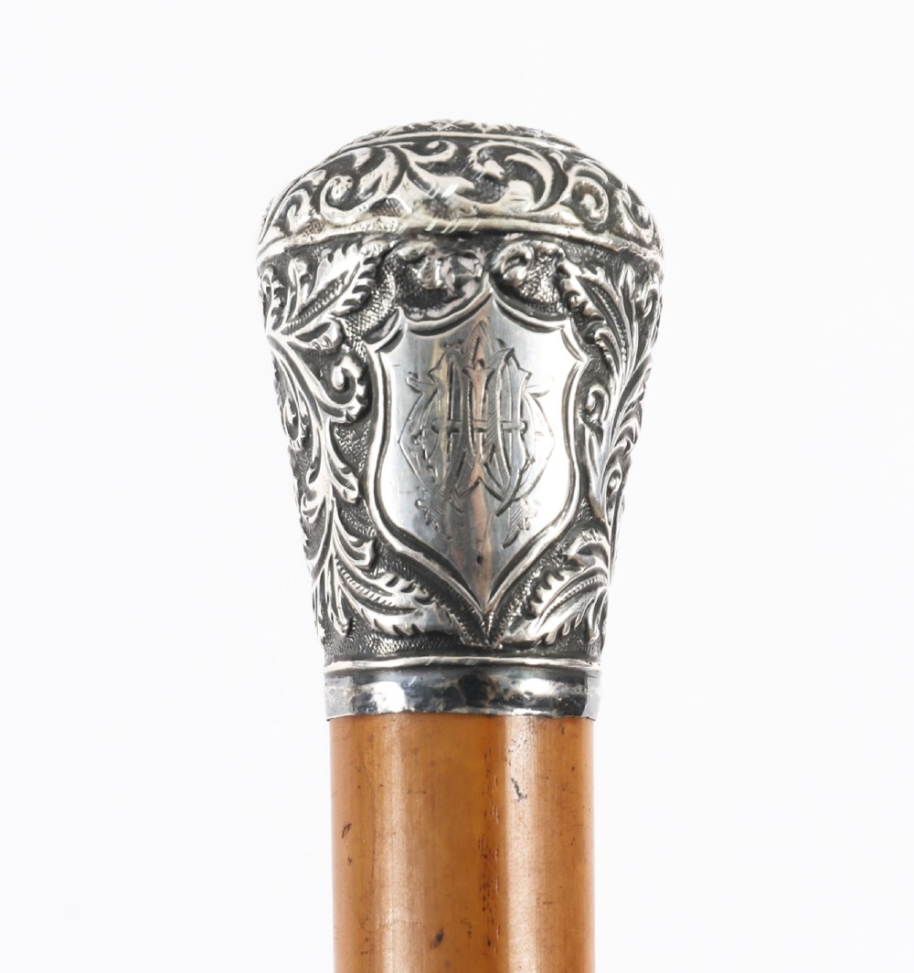 An antique walking stick featuring an elaborately chased silver pommel handle with foliate motifs mounted on a smooth light timber shaft. View V