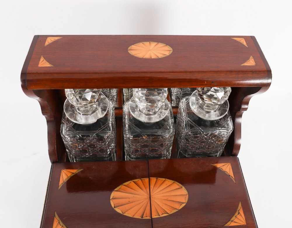 A 19th-century inlaid satinwood and mahogany tantalus containing three cut crystal decanters with faceted stoppers, a mirrored back interior, and a lower fold-out compartment with oval marquetry paterae. View V