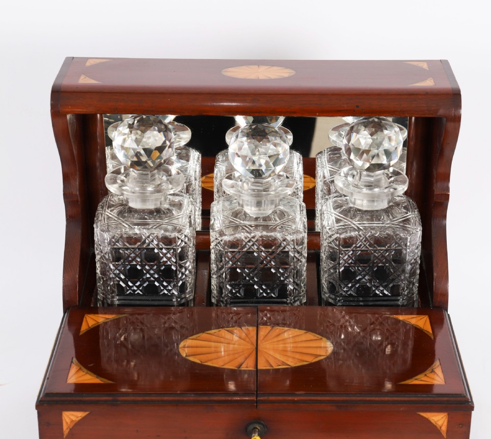 A 19th-century inlaid satinwood and mahogany tantalus containing three cut crystal decanters with faceted stoppers, a mirrored back interior, and a lower fold-out compartment with oval marquetry paterae. View VI