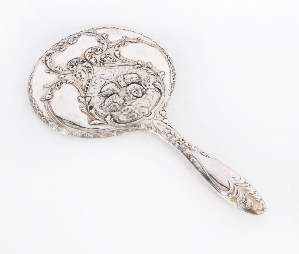An Edwardian sterling silver hand mirror decorated with high-relief repousse cherubs amidst clouds and scrollwork.