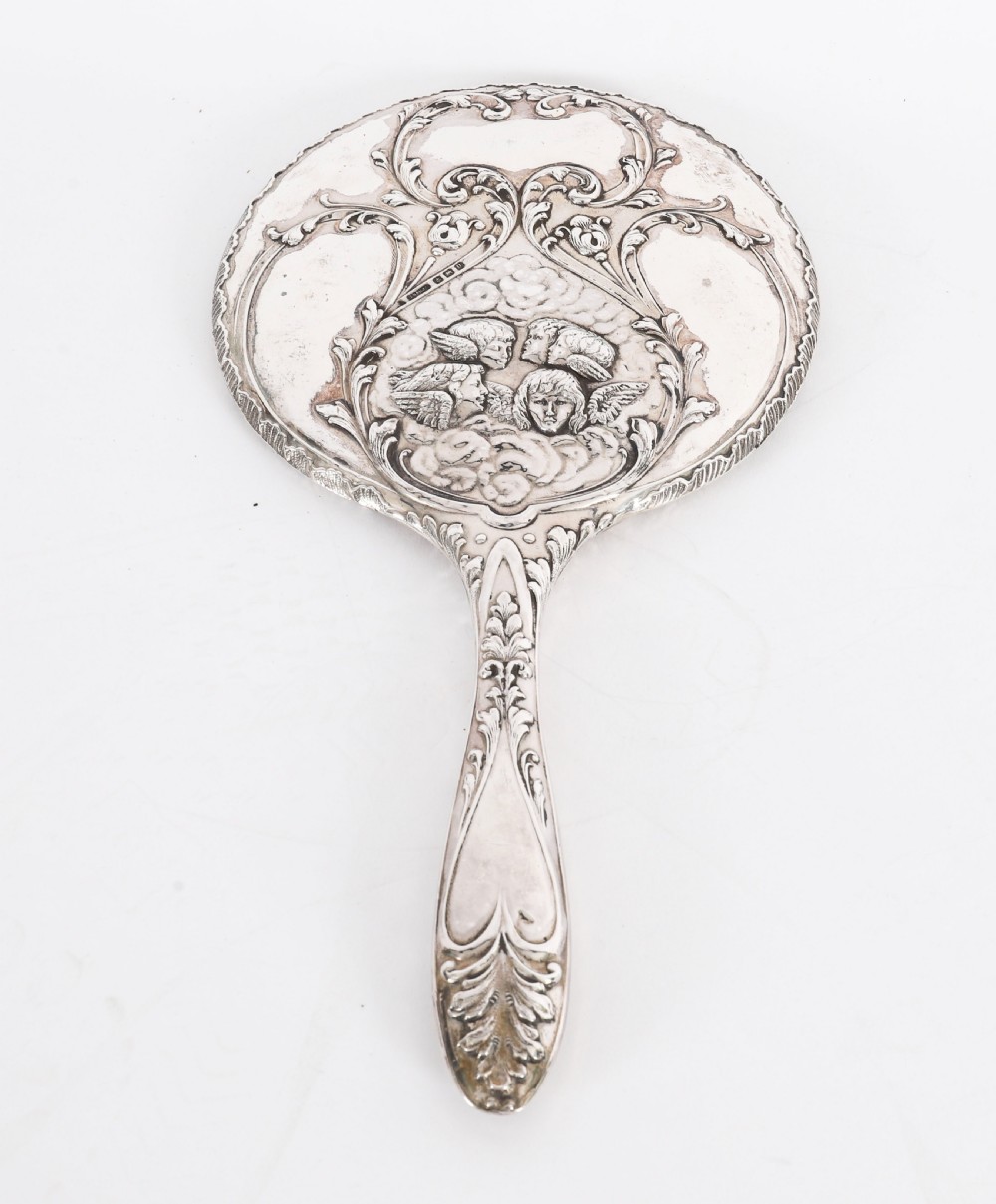 An Edwardian sterling silver hand mirror decorated with high-relief repousse cherubs amidst clouds and scrollwork. View II