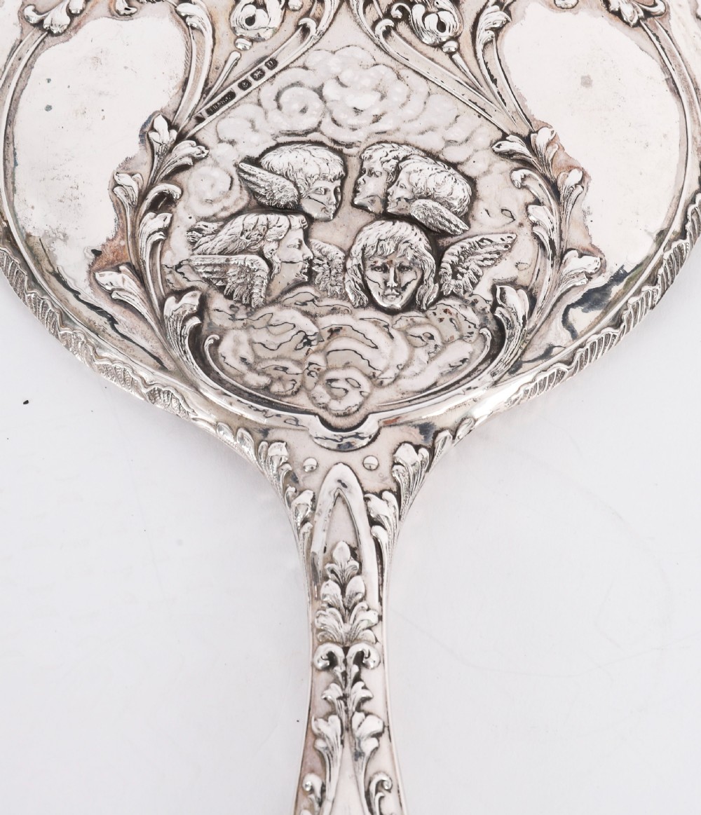 An Edwardian sterling silver hand mirror decorated with high-relief repousse cherubs amidst clouds and scrollwork. View III