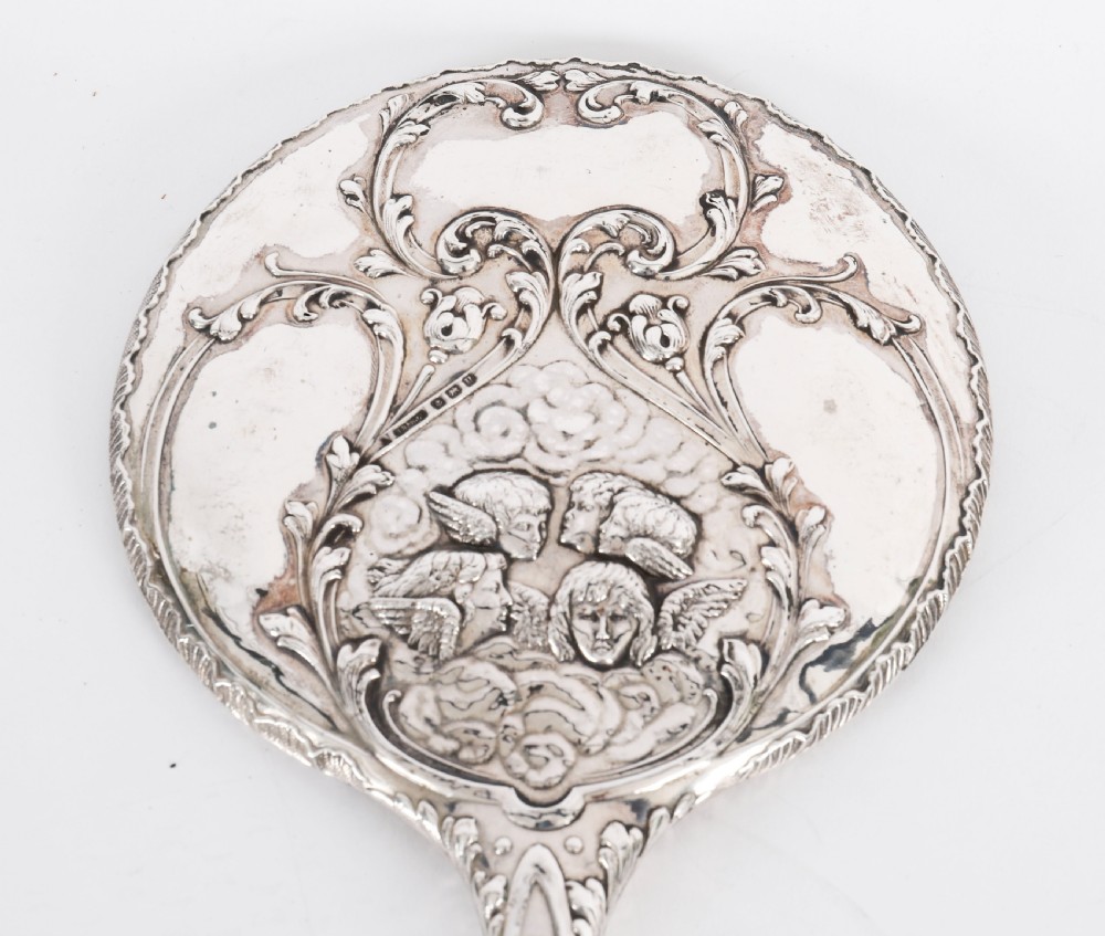 An Edwardian sterling silver hand mirror decorated with high-relief repousse cherubs amidst clouds and scrollwork. View IV