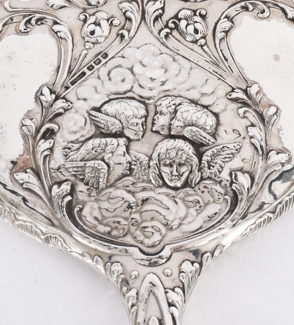 An Edwardian sterling silver hand mirror decorated with high-relief repousse cherubs amidst clouds and scrollwork. View VII