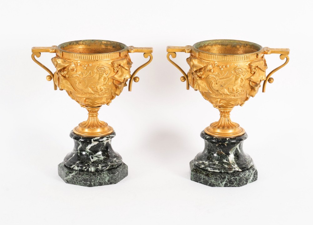 A pair of nineteenth-century French gilt bronze classical urns featuring relief carvings and scroll handles, mounted on dark green marble bases.